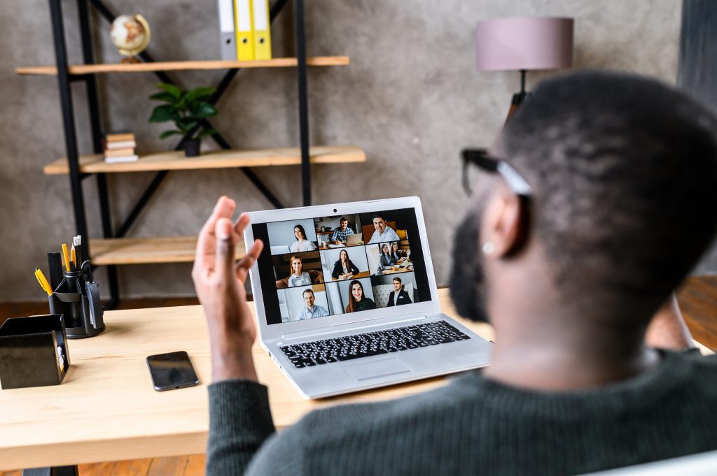 Confident male worker talking online with coworkers, back view, speaks and gestures to many people on video screen. Remote work, virtual meeting virtual event conference