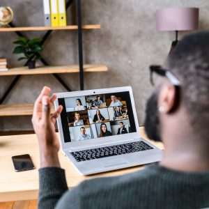 Confident male worker talking online with coworkers, back view, speaks and gestures to many people on video screen. Remote work, virtual meeting virtual event conference