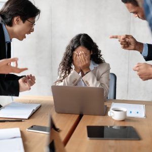 Workplace Conflict And Bullying. Aggressive Coworkers Yelling At Victimized Businesswoman Sitting At Desk In Modern Office. Corporate Communication Problem, Quarrels And Bad Attitude At Work