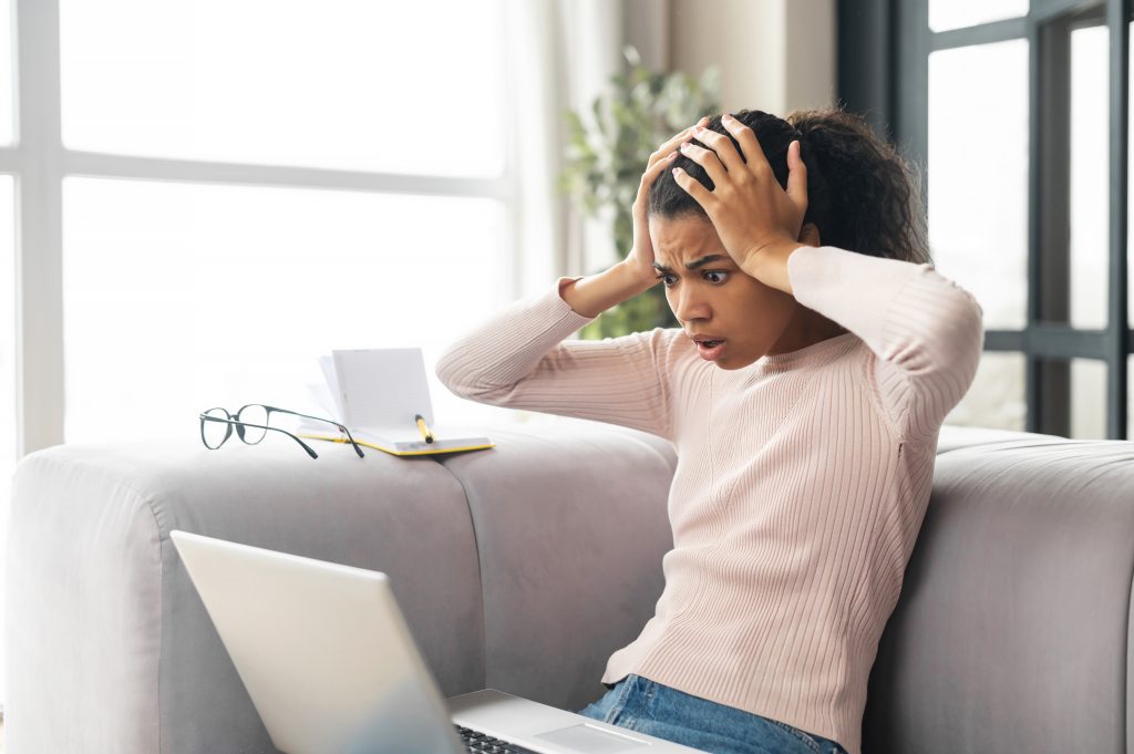 Frustrated young mixed-race female freelancer or student with Afro hairstyle holding head in hands, sitting on the couch with laptop on her lap, stressing out, cannot fix computer issue or a problem stressed mental health wellness wellbeing