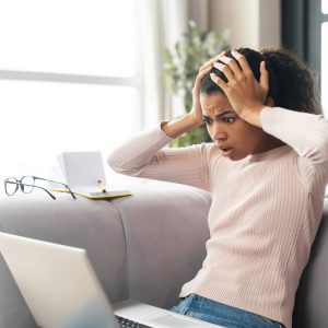 Frustrated young mixed-race female freelancer or student with Afro hairstyle holding head in hands, sitting on the couch with laptop on her lap, stressing out, cannot fix computer issue or a problem stressed mental health wellness wellbeing