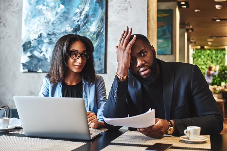 African american businessman and businesswoman are having lunch at restaurant, sitting at table near window, drinking coffee, discussing project, looking through working papers. Man is tired, holding hand on his head, while woman is looking at him with a smile manager management