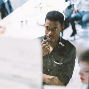 Pensive young boss in formal wear looking at project of employees and analyzing business strategy of developing startup standing in modern office during briefing with colleagues