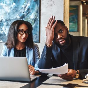 African american businessman and businesswoman are having lunch at restaurant, sitting at table near window, drinking coffee, discussing project, looking through working papers. Man is tired, holding hand on his head, while woman is looking at him with a smile manager management