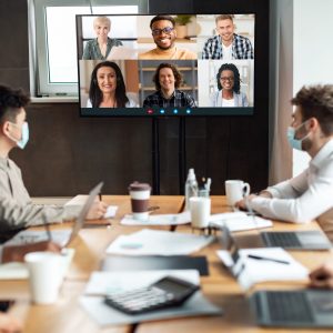 Distant Virtual Meeting Group of diverse business people in masks making online videocall with colleagues, sitting in boardroom, looking at tv screen, talking on web with remote workers