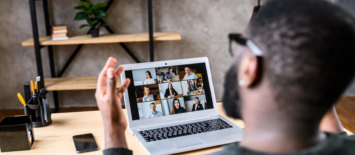 Confident male worker talking online with coworkers, back view, speaks and gestures to many people on video screen. Remote work, virtual meeting virtual event conference