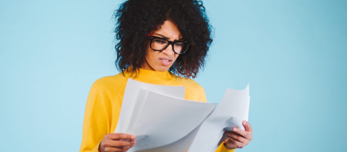 Worried african american businesswoman reading a notification standing on blue background in yellow wear and afro hairstyle.