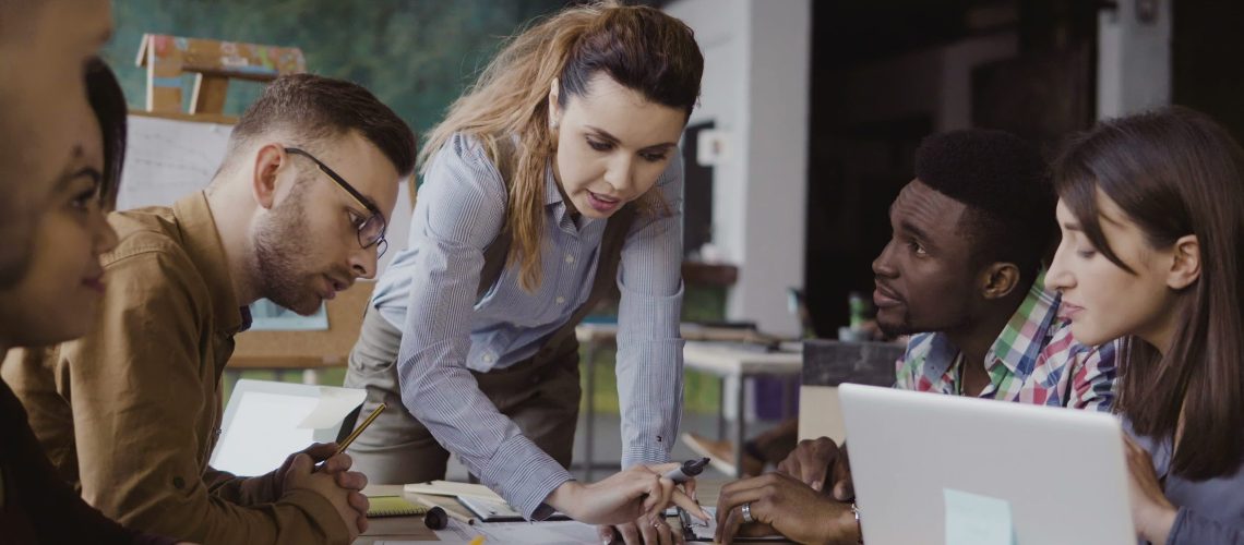 Brunette female team leader talking with mixed race group of people, writes with a marker on the model of house. Brainstorming of start-up company in modern office. Creative architectural project.