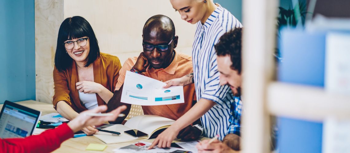 Diverse group of young people dressed in smart casual wear discussing graphics and sketches during brainstorming in office.Modern hipster students teamworking on common task sitting at meeting table