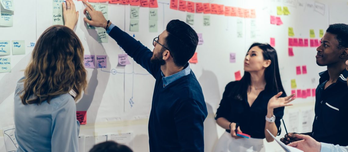 Team of multicultural young people pointing on wall with glued colorful paper notes with foreign words during productive lesson.Diverse group of male and female employees in formal wear using stickers