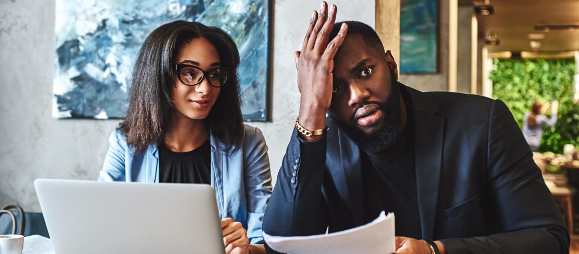 African american businessman and businesswoman are having lunch at restaurant, sitting at table near window, drinking coffee, discussing project, looking through working papers. Man is tired, holding hand on his head, while woman is looking at him with a smile manager management