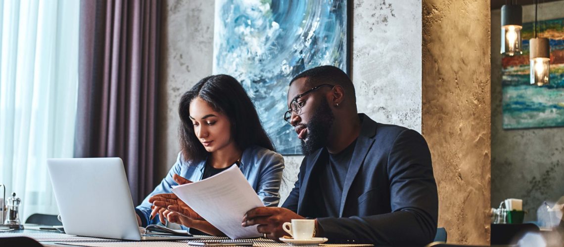 African american businessman and businesswoman having lunch at restaurant, sitting at table near window, drinking coffee, discussing project. Man explains his ideas, looking at the screen of laptop