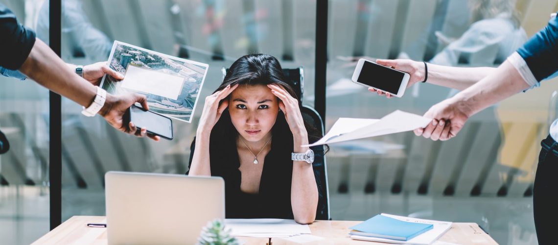 Young disappointed woman feeling headache from deadline surrounded by colleagues at workplace, multi-purpose female employee,tired of work and exhausted, stress in office. Overworked female stressed unhappy battling to cope mental health wellness wellbeing overworked