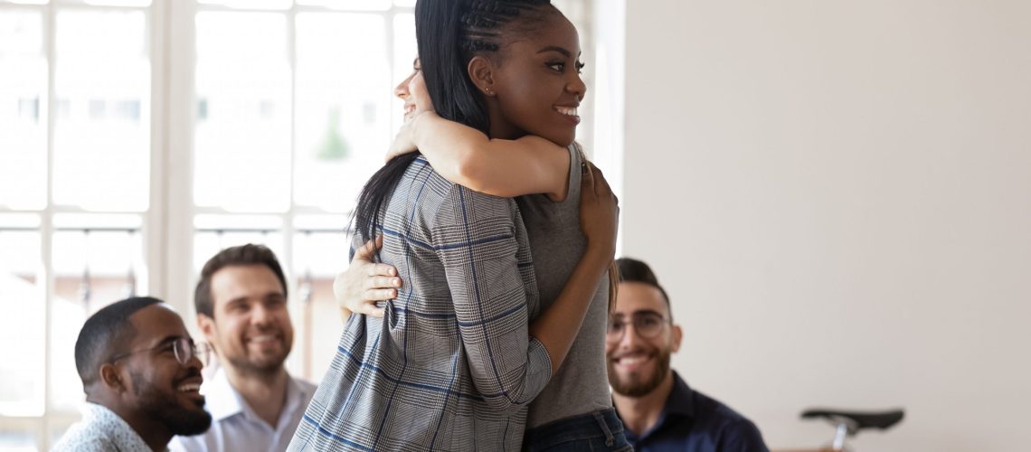 Happy african american manager hugging smiling female colleague at team building activity. Two friendly coworkers embracing at group therapy session. Joyful diverse team supporting each other.