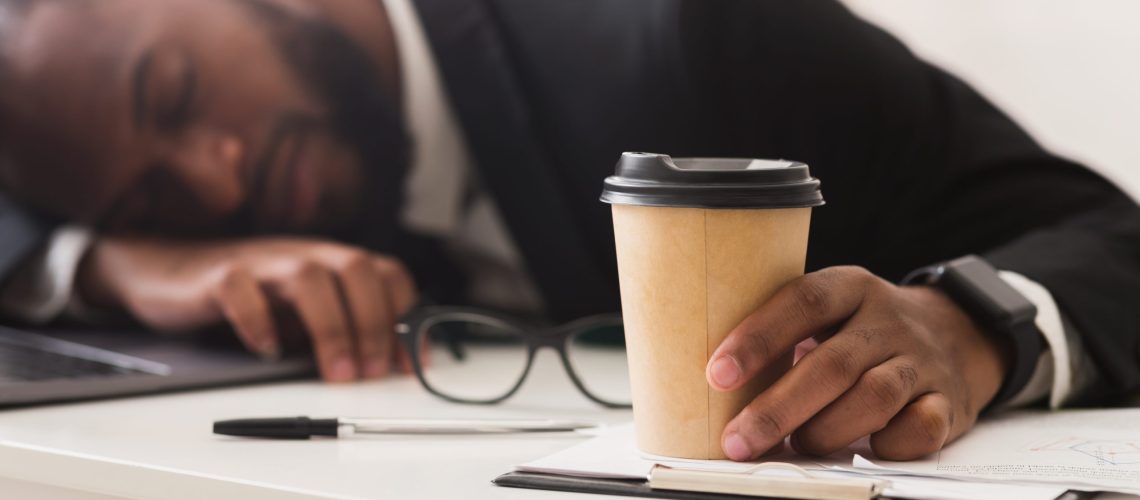 Exhausted african business clerk holding cup of coffee and sleeping at workplace, burnout concept