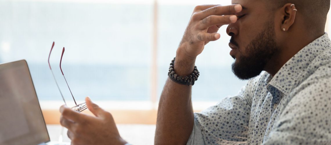 Close up side view african man sitting at desk near computer take break from study or work took off glasses rub nose bridge to reducing eye strain. Dry eyes or poor vision caused by pc overuse concept