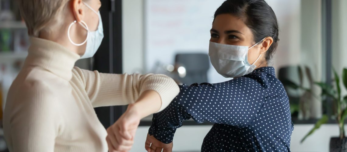 Smiling diverse female colleagues wearing protective face masks greeting bumping elbows at workplace, woman coworkers in facial covers protect from COVID-19 coronavirus in office, healthcare concept health wellness in the workplace wellbeing clean work environment office