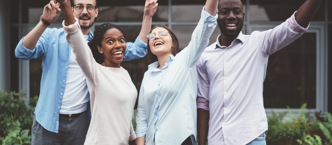 Optimistic multiethnic young people in casual outfits smiling and raising arms while standing in garden of suburban house during meeting