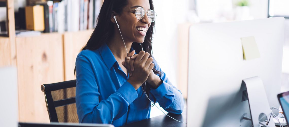 Happy young African American female office worker in blue shirt and glasses sitting at work table with  hands together wearing headphone and conducting online negotiations with coworkers