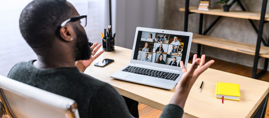 Virtual meeting with many people together. African-American young guy talking online with employees via video connection. Multiracial team. Back view