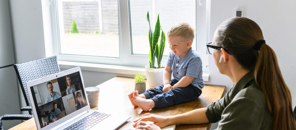 Stay at home. Mother working from home, she talks online with a coworkers via video call on laptop, a cute kid near