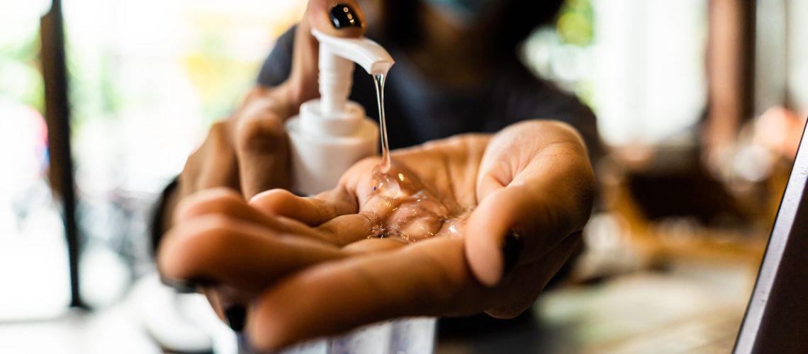 Young woman washing hands with alcohol gel in quarantine for coronavirus wearing protective mask with social distancing and using laptop computer working at home. Work from home and covid-19 concept.