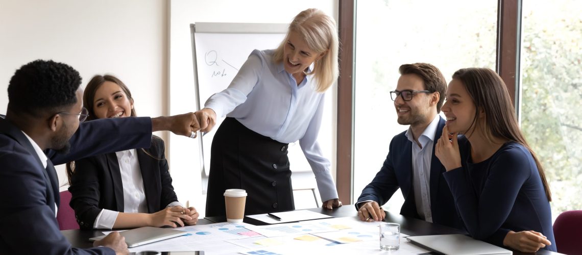 Mature businesswoman bumping fists with young black male colleague excited by success of their common project, friendly senior female employee jokingly showing respect to millennial african teammate