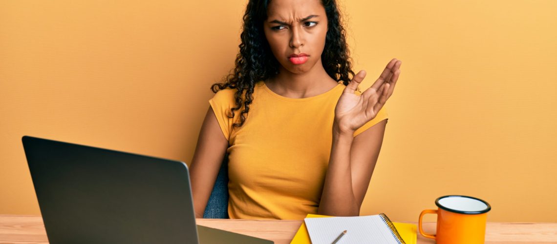 Young african american girl doing video call waving to laptop clueless and confused expression. doubt concept.