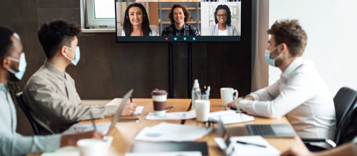 Distant Virtual Meeting Group of diverse business people in masks making online videocall with colleagues, sitting in boardroom, looking at tv screen, talking on web with remote workers