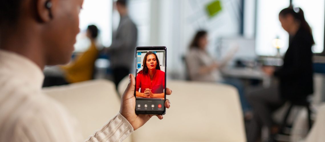 Over the shoulder of employee discussing with team member on video call using phone in start up business office. woman during video meeting, online conference planning new financial project