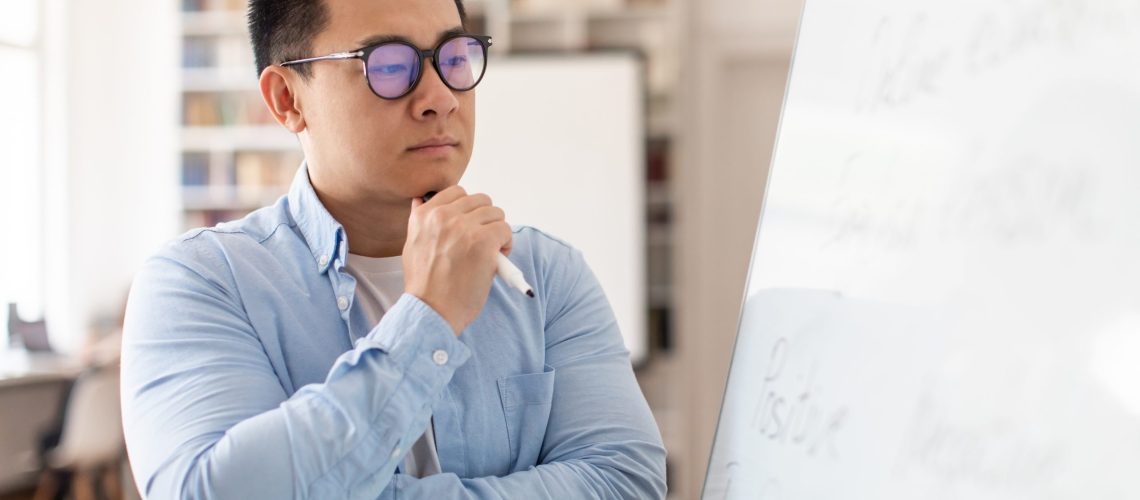 Thoughtful Asian Middle Aged Man Teacher Thinking Looking At Whiteboard Having Class Standing In Modern Classroom Indoors, Wearing Eyeglasses. Education And Science Concept. Selective Focus