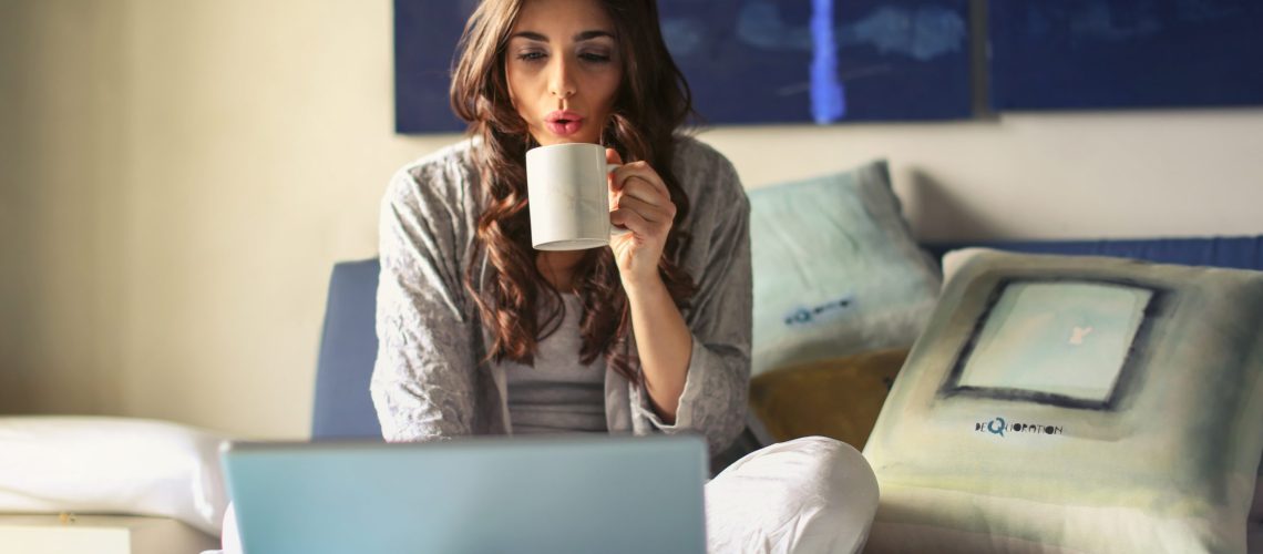 woman-in-grey-jacket-sits-on-bed-uses-grey-laptop-935743