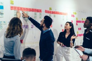 Team of multicultural young people pointing on wall with glued colorful paper notes with foreign words during productive lesson.Diverse group of male and female employees in formal wear using stickers