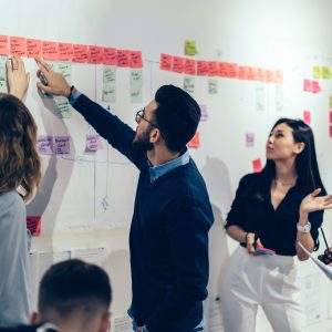 Team of multicultural young people pointing on wall with glued colorful paper notes with foreign words during productive lesson.Diverse group of male and female employees in formal wear using stickers