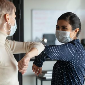 Smiling diverse female colleagues wearing protective face masks greeting bumping elbows at workplace, woman coworkers in facial covers protect from COVID-19 coronavirus in office, healthcare concept health wellness in the workplace wellbeing clean work environment office